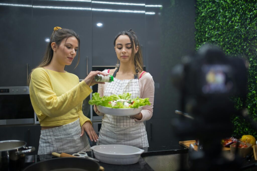 Two women preparing a salad while recording a cooking video in a stylish kitchen setting.